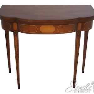 May include: A dark brown, half-moon shaped antique table with a decorative inlaid design. The table has four slender, tapered legs and a scalloped edge. The table's drawer features a rectangular inlay. The table is approximately 30 inches wide.