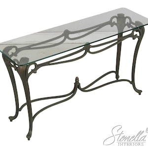 May include: A rectangular console table featuring a clear glass top and a dark bronze-toned metal base. The base is constructed with curved, flowing metal supports that connect the four legs, creating an open frame design with a central finial.
