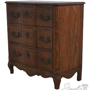 May include: A dark brown wooden dresser with three drawers, each with ornate metal handles. The dresser has a curved front and legs, and a dark top. The wood grain is visible throughout the piece, giving it a rustic look.