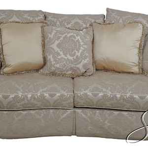 May include: A cream-colored, curved sofa with a damask pattern upholstery. The sofa has three pillows with a cream-colored fabric and a fringe trim.