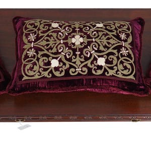May include: Three deep burgundy velvet throw pillows arranged on a dark wood table. Two square pillows feature centered gold-toned embroidery, while the central rectangular pillow displays an intricate gold and white floral-inspired pattern.