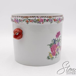May include: White ceramic planter with a floral design and a red face accent. The planter features a colorful floral border and a cluster of pink, purple, and blue flowers. A decorative piece for home decor.