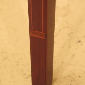Pu&ograve; includere: Primo piano di una gamba di tavolo in mogano con strisce di legno intarsiate. La gamba &egrave; quadrata e ha una finitura marrone rossiccio scuro.