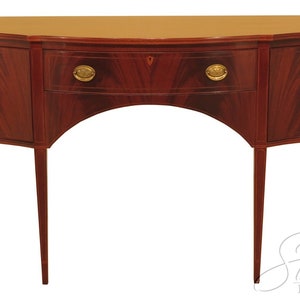 Pu&ograve; includere: Un credenza in legno di mogano con due ante e un cassetto. La credenza ha un fronte curvo e poggia su quattro gambe sottili.