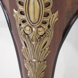 May include: Close-up of a wooden table leg with a gold metal decorative accent. The accent features a detailed floral design and a central oval shape.