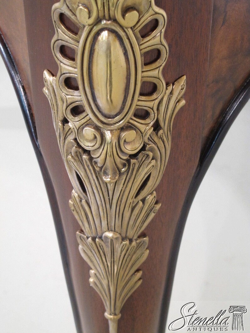 May include: Close-up of a wooden table leg with a gold metal decorative accent. The accent features a detailed floral design and a central oval shape.