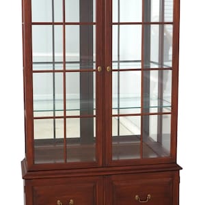 May include: A tall, dark cherry wood display cabinet featuring two glass-paned doors with brass knobs, revealing interior glass shelving. The base includes two wooden drawers with brass pulls, all supported by a sturdy, bracket-style footed frame.