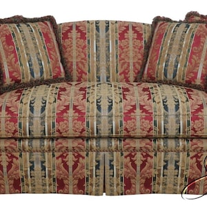 May include: A patterned sofa with two matching throw pillows. The sofa features a red, gold, and beige floral design with a skirted base. The pillows have a striped pattern with a brown fringe. The sofa has curved arms and a high back.