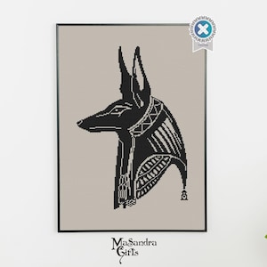 May include: Black and white cross-stitch art of Anubis, the Egyptian god of the afterlife, in a framed print. The artwork features intricate details and is set against a neutral background. The Masandra Girls logo is at the bottom.