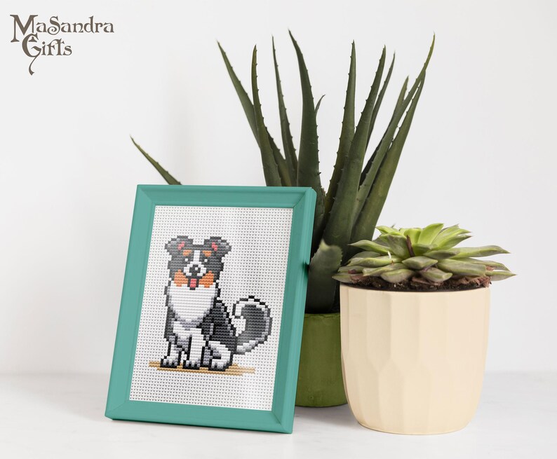 A vibrant, teal-framed cross-stitch artwork of a sitting dog, featuring black, brown, and white colors, displayed against a bright white background. Nearby, there are two potted plantsa tall, green aloe vera plant and a succulent in a beige geometric pot. The image conveys a cozy and artistic home decor vibe, perfect for dog lovers and plant enthusiasts. Ideal for gift ideas or interior design inspiration, this arrangement combines the charm of handcrafted art with nature's beauty.