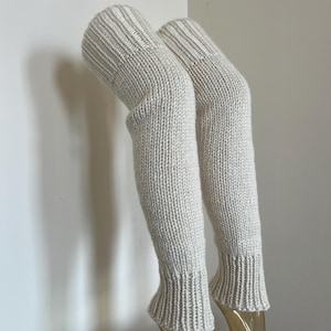 May include: A pair of off-white, knee-high leg warmers featuring a thick, chunky knit texture. The leg warmers have a ribbed cuff at the top and bottom, providing a snug fit. The knit pattern is consistent throughout the length of the garment.