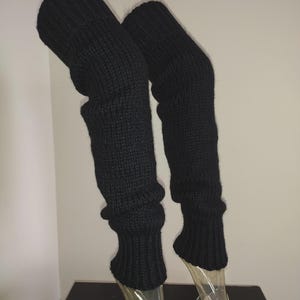 Op de afbeelding: Een paar lange, zwarte gebreide beenwarmers met een geribbelde textuur over de gehele lengte. De beenwarmers worden getoond op doorzichtige plastic vormen, met een dikke, elastische boord aan de bovenkant en een bijpassende geribbelde boord onderaan.