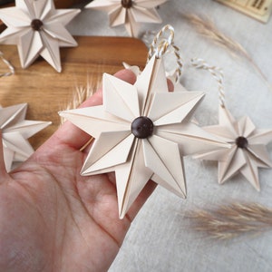 May include: A set of six beige paper star ornaments with brown button centers. The stars are strung together with twine and are hanging from a wooden surface.