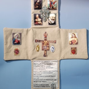 Peut inclure: Une tenture murale en tissu beige en forme de croix ornée d'images religieuses, d'un crucifix central et de médailles en métal. La section inférieure comporte un texte italien imprimé commençant par 'Altola! Fermati, demonio' et finissant par 'Confido e spero in te'.