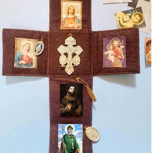 May include: A brown felt cross-shaped religious item with images of Jesus, Mary, and other religious figures. Includes a metal crucifix, a gold-coloured clasp, and a safety pin.