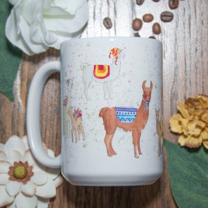 May include: White ceramic mug with a colorful illustration of llamas wearing hats and saddles. The mug has a white background with a light blue rim.