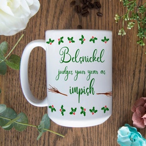 May include: White ceramic coffee mug with a green and red holly border. The mug has a green inscription that reads "Belsnickel judges your year as impish" with a brown twig design.