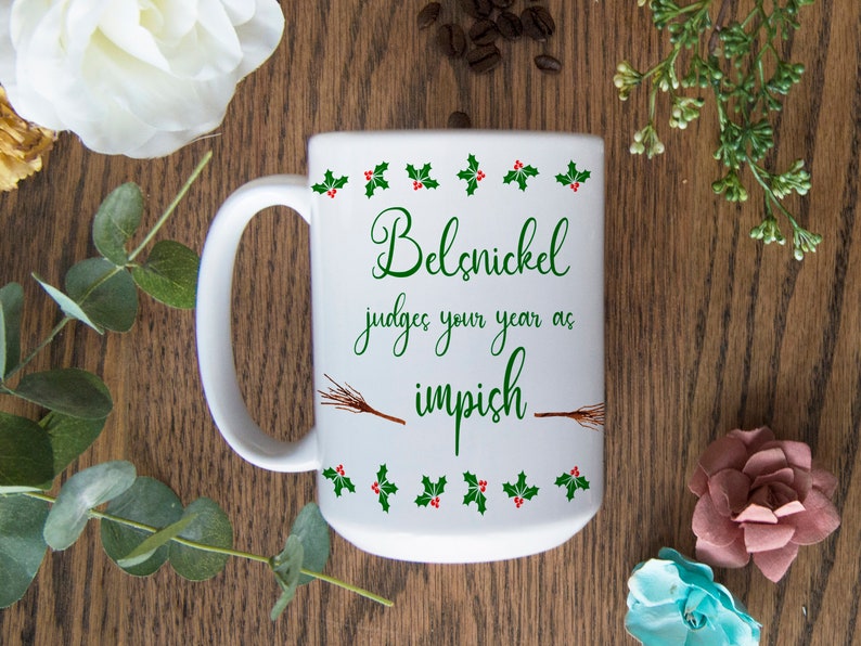 May include: White ceramic coffee mug with a green and red holly border. The mug has a green inscription that reads "Belsnickel judges your year as impish" with a brown twig design.