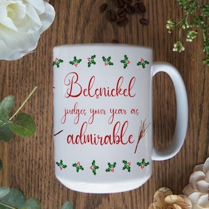 May include: White ceramic mug with a red and green design. The mug has a handle and says "Belsnickel judges your year as admirable" in red cursive text.
