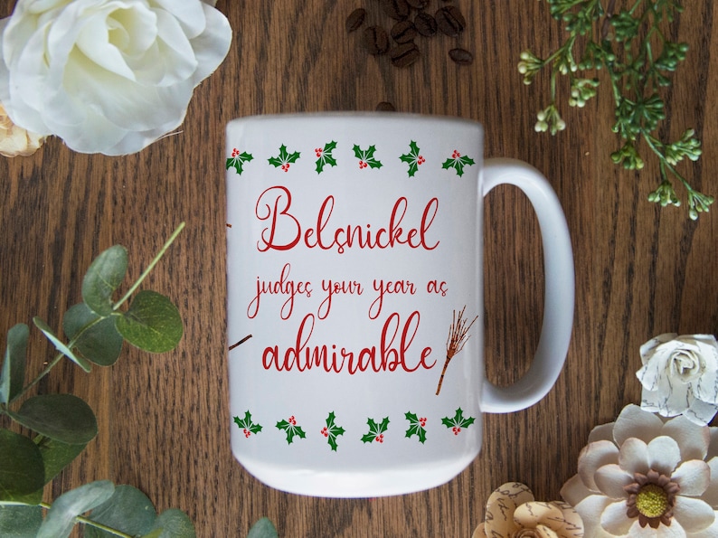 May include: White ceramic mug with a red and green design. The mug has a handle and says "Belsnickel judges your year as admirable" in red cursive text.