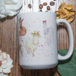 May include: White ceramic mug with a colorful illustration of four llamas wearing hats and accessories. The llamas are surrounded by pink and white splatters.