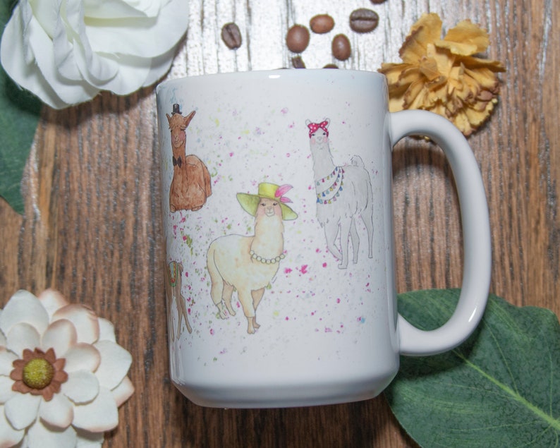 May include: White ceramic mug with a colorful illustration of four llamas wearing hats and accessories. The llamas are surrounded by pink and white splatters.