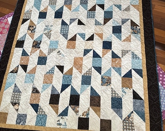 May include: A quilt with a geometric pattern in shades of blue, brown, and beige. The quilt is made of various fabrics with different patterns and textures. The quilt is edged with a black border and a gold trim.