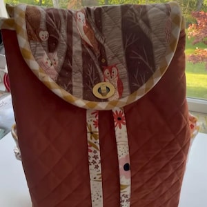 May include: Quilted backpack in a rust-orange color with a flap featuring an owl and tree design. The flap is trimmed with a yellow and white checkered pattern. The straps are white with a floral pattern and the closure is a gold-tone clasp.
