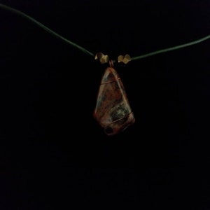 May include: A pendant necklace featuring a triangular, multi-colored stone wrapped in copper wire. The stone displays shades of brown, green, and red. The necklace has a green cord with gold-colored beads near the pendant.