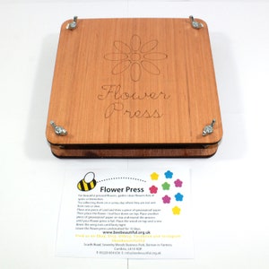 May include: Wooden flower press with a daisy design and the words "Flower Press" engraved on the top. The press has four metal wing nuts and a printed card with instructions and a logo.