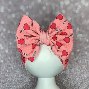 May include: A pink baby headband with a repeating pattern of red heart-shaped lollipops. The bow is tied in the center and sits atop a white, distressed pedestal. The background is a sparkly, silver bokeh.