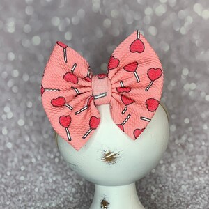 May include: A pink bow with a white lollipop and heart pattern. The bow is on a white pedestal.