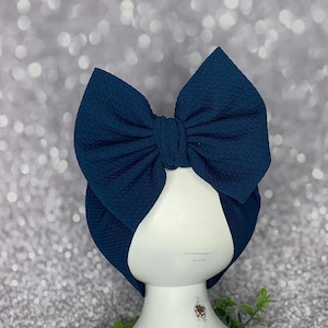May include: A navy blue baby bow headband. The bow is large and textured, with a matching navy blue band. The headband is displayed on a white, distressed pedestal, set against a sparkly, silver background. The bow is a stylish accessory.