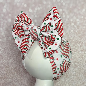 May include: A white headband with a large bow featuring a Christmas tree pattern in red and green. The bow is centered on a white mannequin head, with a glittery background.