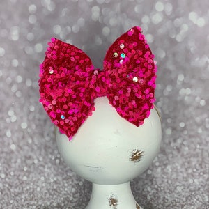 May include: A bright pink sequined hair bow. The bow is atop a white, distressed wooden pedestal. The background is a soft, blurred silver and white. The bow is a vibrant, eye-catching accessory.