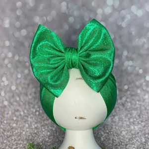 May include: A vibrant green bow headband. The bow is made of a shiny, textured fabric and is centered on a matching green headband. The headband is displayed on a white, distressed pedestal, set against a sparkly background.