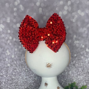 May include: Red sequin bow headband on a white mannequin head. The headband is on a white stand with a white base. The stand is on a woven wicker surface.