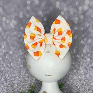 May include: A large, white hair bow with a candy corn pattern. The bow is made of fabric and is attached to a thin, yellow headband. The bow is displayed on a white, distressed pedestal.
