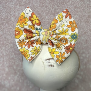 May include: A large, fabric hair bow featuring a retro floral pattern in shades of yellow, orange, and brown on a white background. The bow is mounted on a white, distressed wooden stand, showcasing its design and style. The bow is a hair accessory.