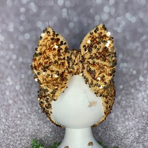 May include: A tan and gold sequined bow on a white mannequin head. The bow is made of a soft, textured fabric and is covered in small, shiny sequins. The mannequin head is on a white pedestal.