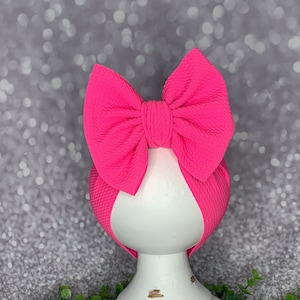May include: A bright pink baby bow headband. The bow is large and textured, and the headband is wrapped around a white, distressed wooden form. The background is a soft, blurred silver. The headband is displayed on a white pedestal.