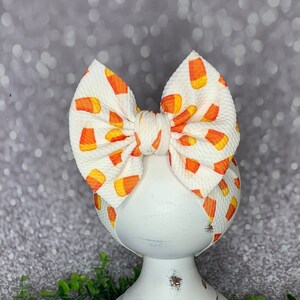 May include: A white baby headband with a large bow featuring a candy corn pattern. The bow is cream-colored with orange and yellow candy corn graphics. The headband is displayed on a white pedestal.