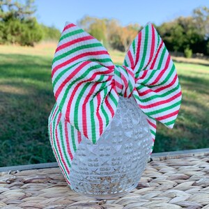 May include: Red and green striped fabric headband with a large bow. The headband is tied in a bow and is sitting on a woven surface.