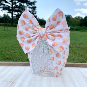 May include: A white baby bow with a repeating pattern of candy corn in orange, yellow, and white. The bow is tied in the center and has frayed edges. The bow is sitting on a clear glass object.