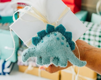 May include: A light blue felt dinosaur ornament with dark blue dorsal plates and stitched details, attached to a white square gift box wrapped with thin cream-colored ribbon and accented with white feathers on top.