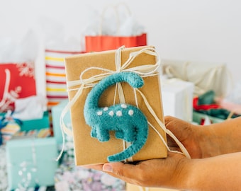 May include: A brown paper gift box tied with thin white string and adorned with a teal felt dinosaur ornament featuring small white polka dots on its back. The dinosaur has a long neck and tail, and is attached to the center of the wrapped package.