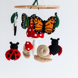 May include: A handmade baby mobile featuring felted insects and plants. The mobile includes a monarch butterfly, ladybugs, mushrooms, a snail, and leaves. The wooden ring at the top has the word "DIVAS" printed on it.
