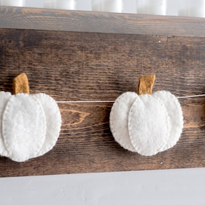 May include: A white felt pumpkin garland with brown felt stems hangs on a dark brown wooden surface.