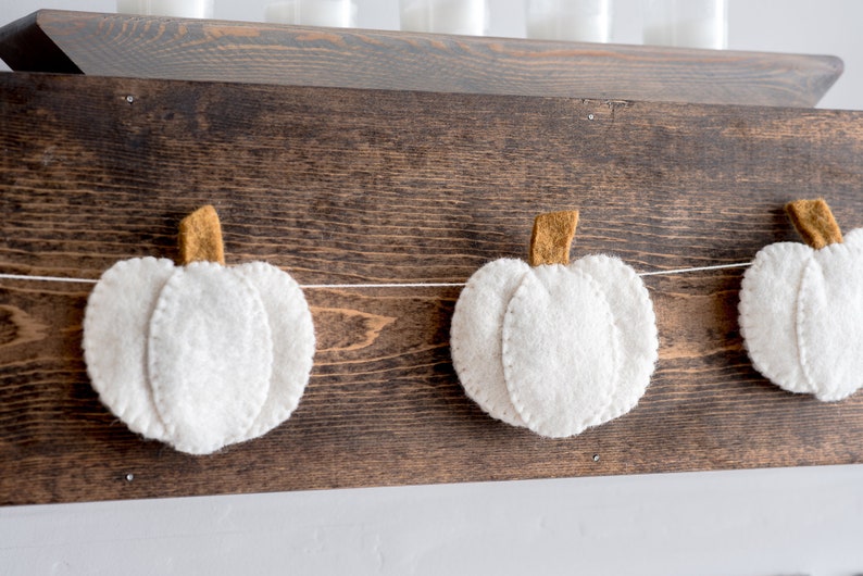 May include: A white felt pumpkin garland with brown felt stems hangs on a dark brown wooden surface.