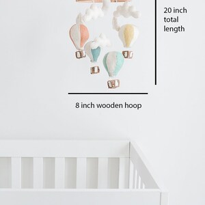 Op de afbeelding: Een baby mobile met kleurrijke vilten luchtballonnen en witte wolkenaccenten. De mobile hangt aan een houten ring, met een totale lengte van 50.8 cm. De houten ring heeft een diameter van 20.3 cm.